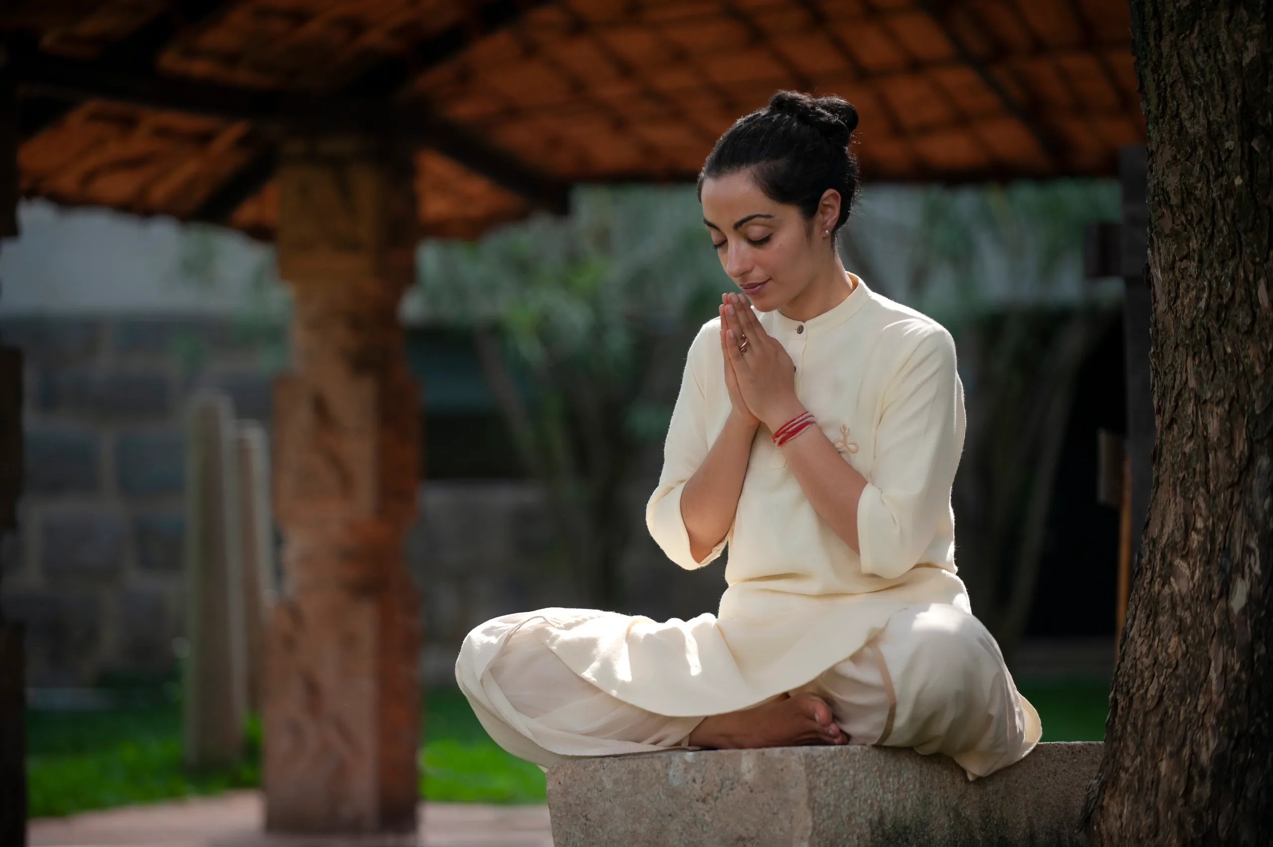A practitioner sits in a quiet gesture of prayer beneath a traditional tiled pavilion.