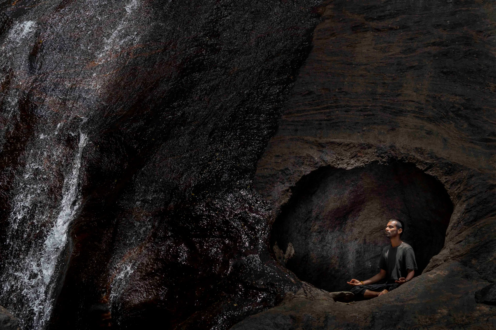 A lone practitioner sits in deep meditation within a rock hollow beside a mountain waterfall.