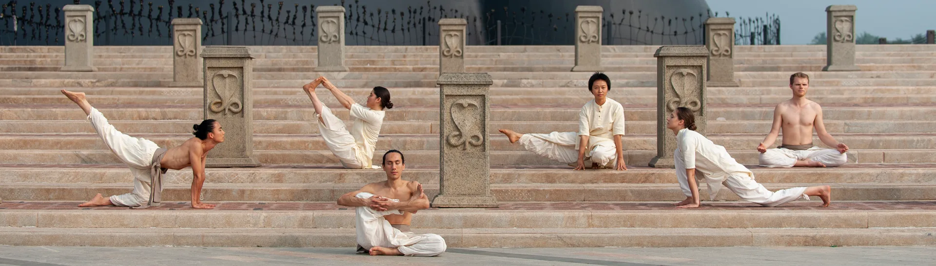 A group of practitioners holding Classical Hatha Yoga postures together on temple steps at dawn.