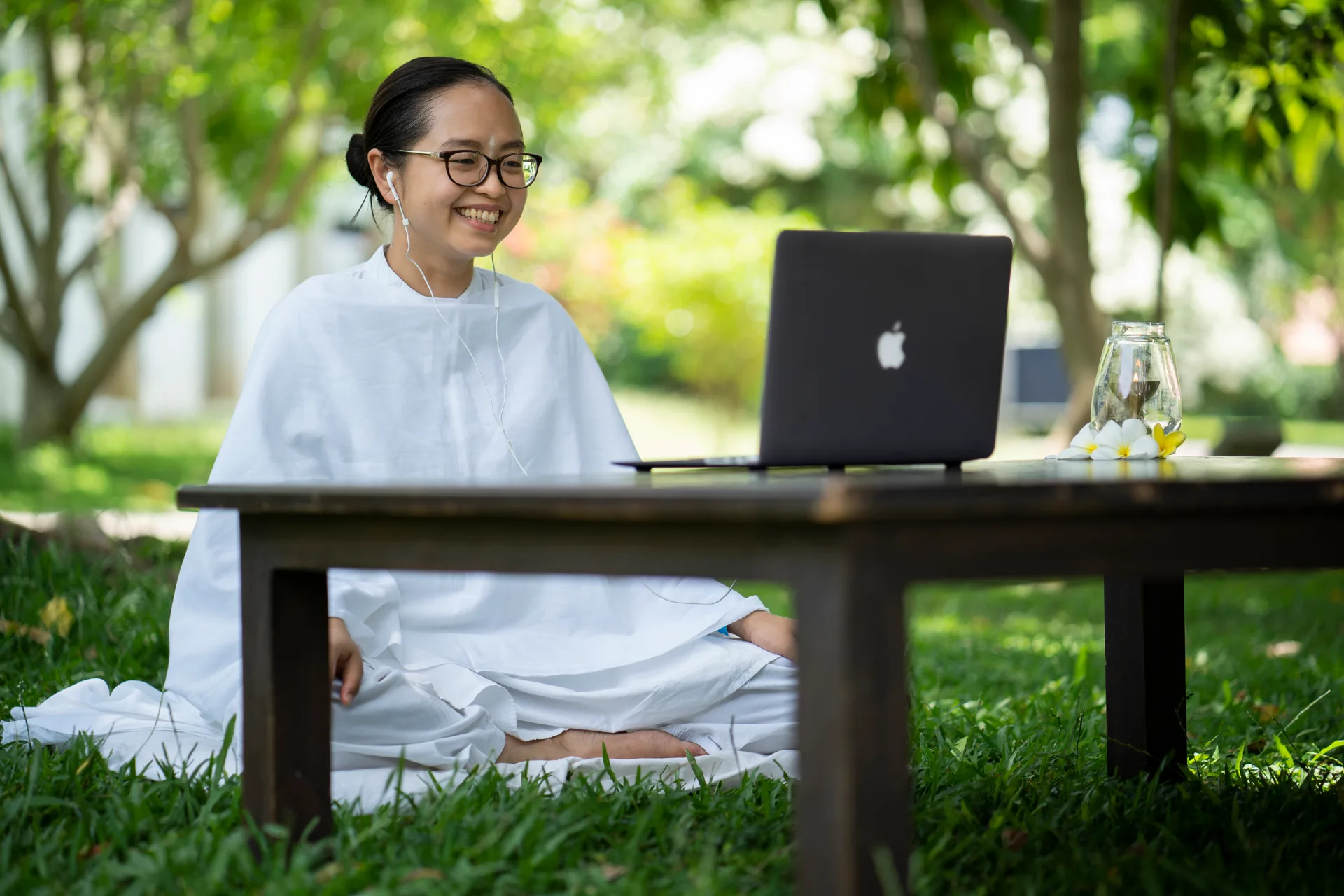 A student in white joins a live online Classical Hatha Yoga session on a laptop, seated in a sunlit garden.
