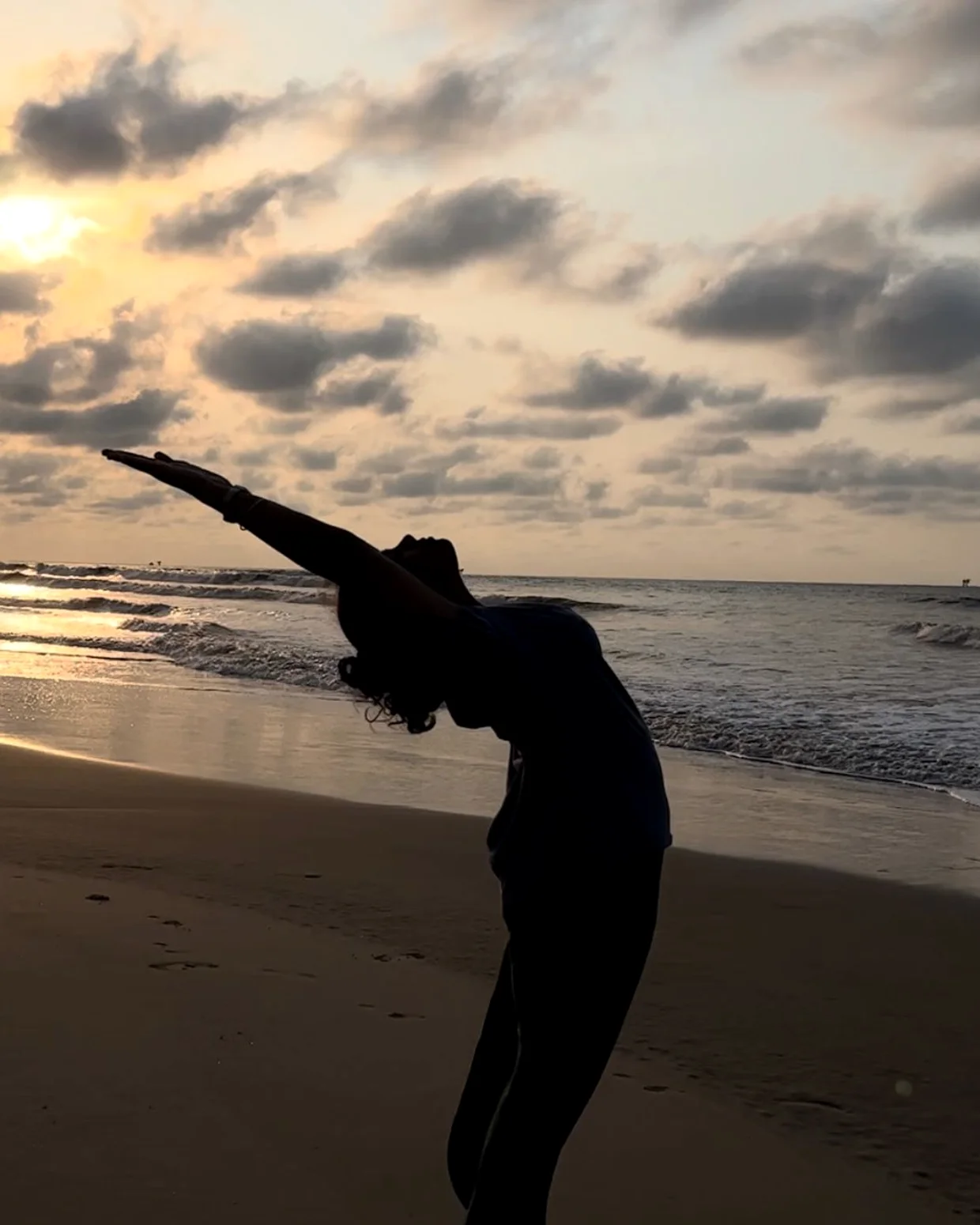 A lone figure arches into a backbend on an empty beach as the sun rises over the water.