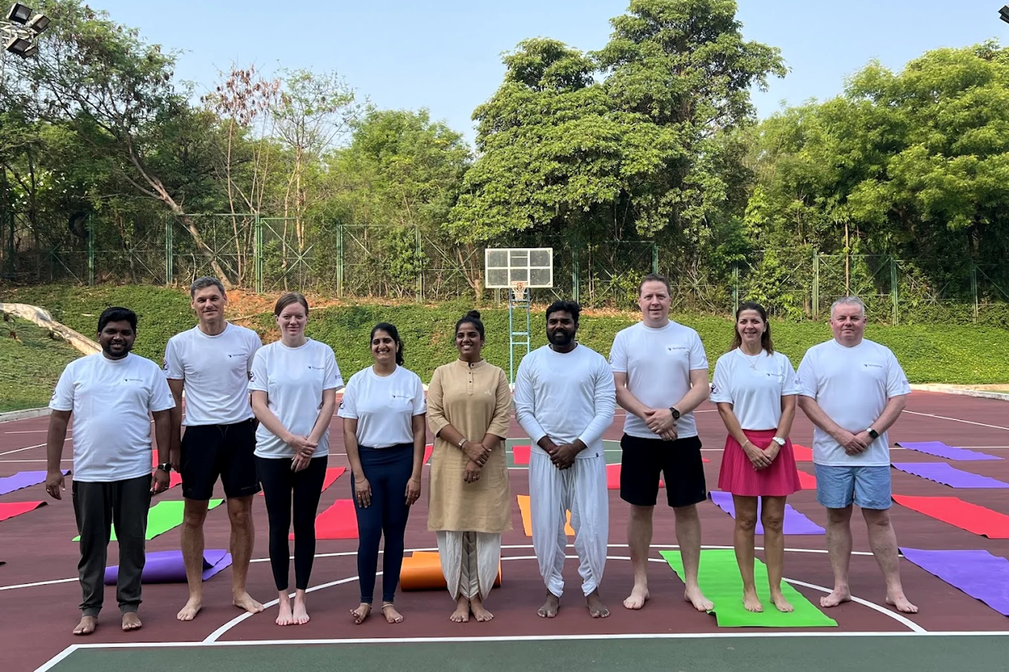 A small international group stands with two Trikonam teachers after a session, mats laid out behind them.