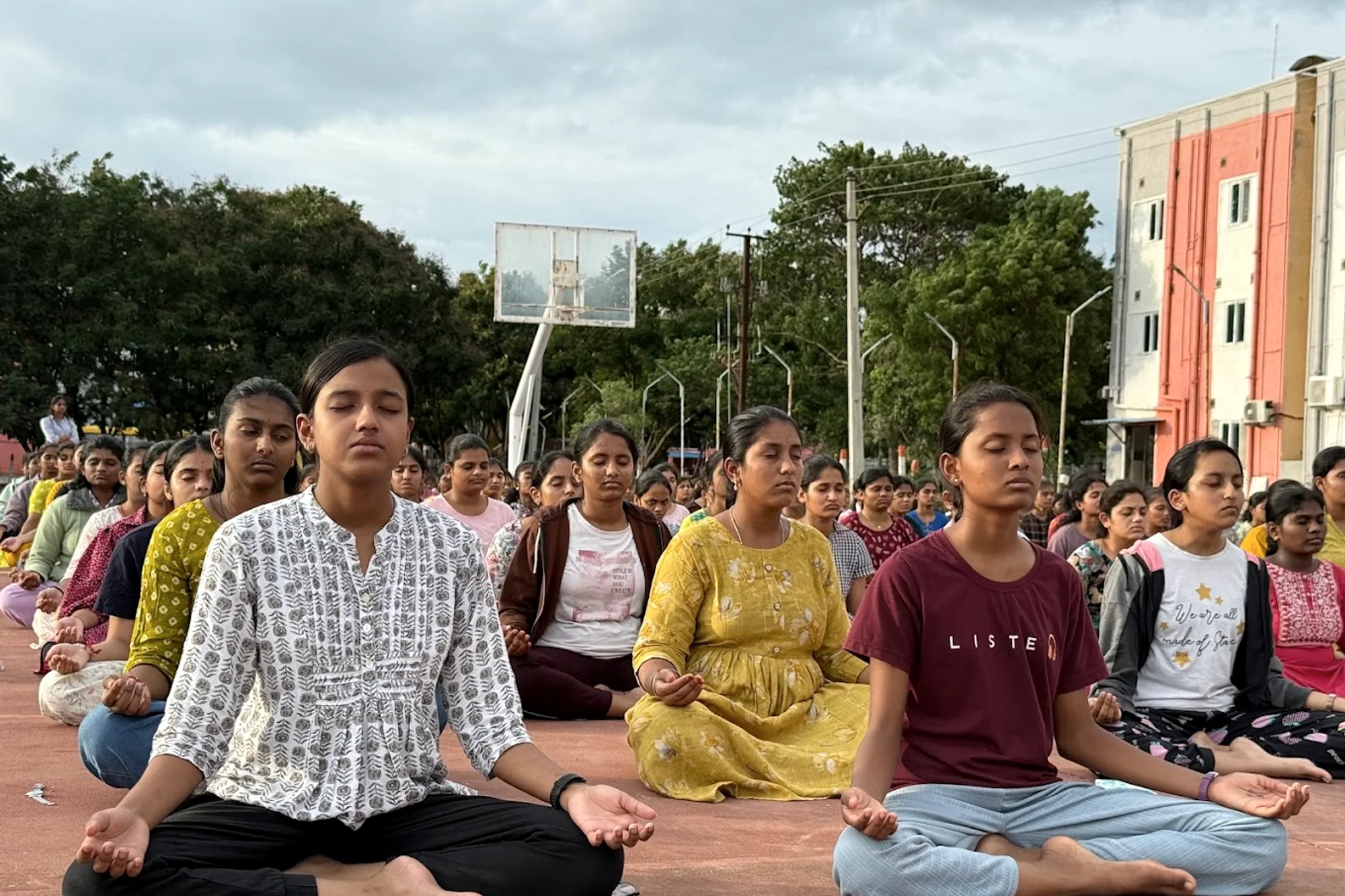 A large group sits in quiet meditation on an open ground, eyes closed, hands resting.