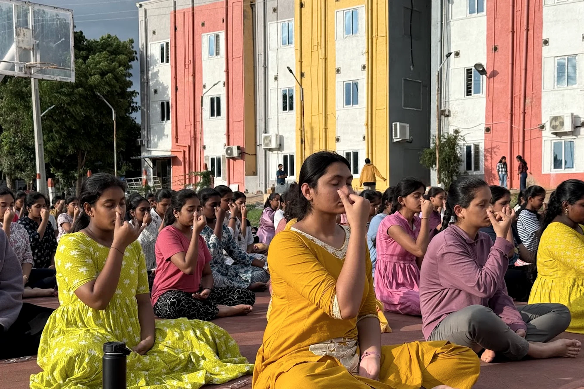 Students seated in rows on an open ground practising alternate-nostril breathing together in the morning light.