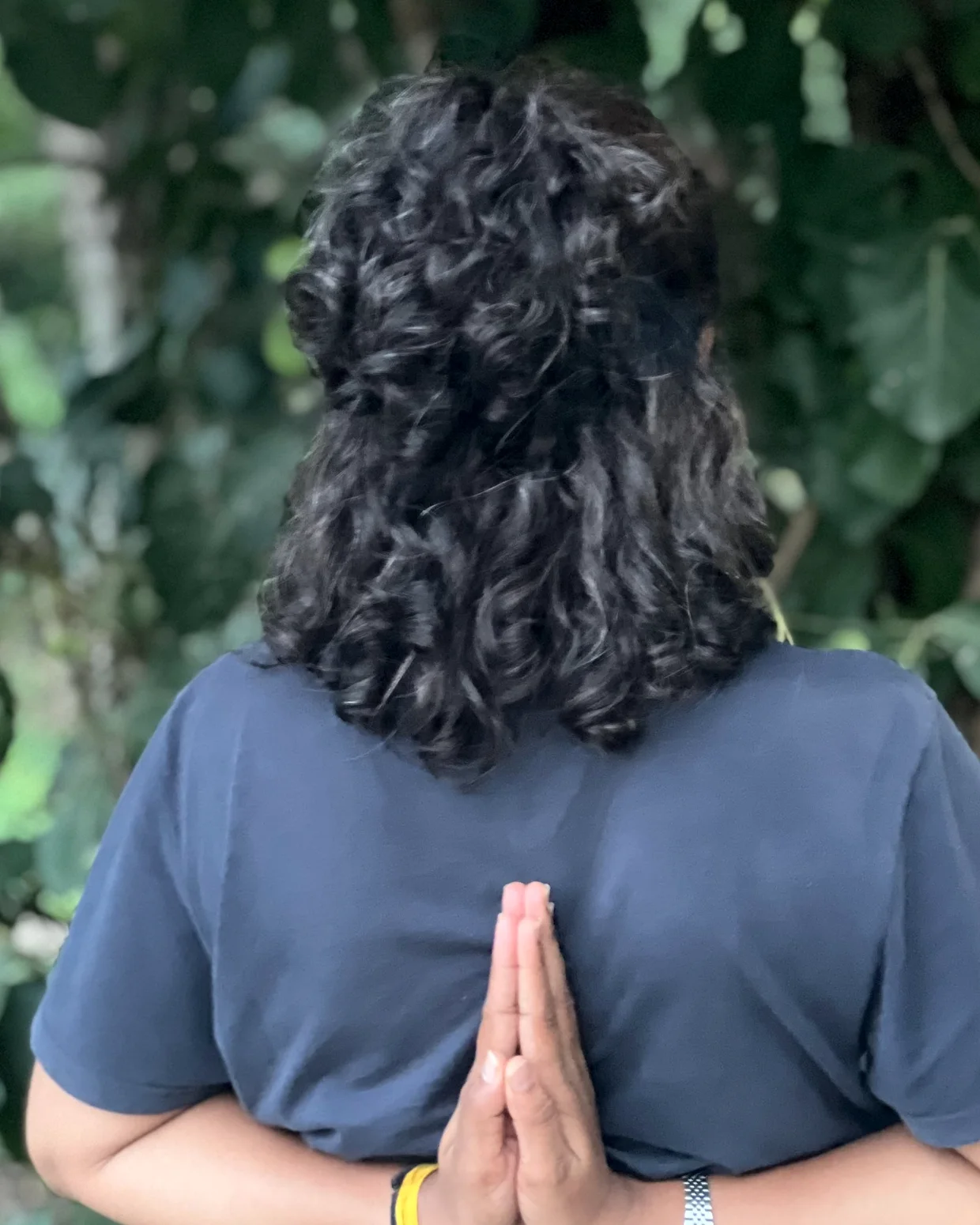 Seen from behind, a practitioner joins the palms in reverse prayer between the shoulder blades, green foliage around.