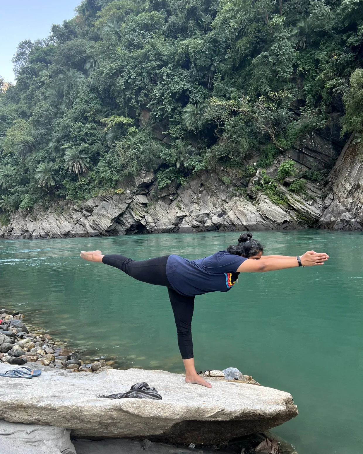 A practitioner holds a standing balance on a flat rock beside a still green river, forested hills behind.