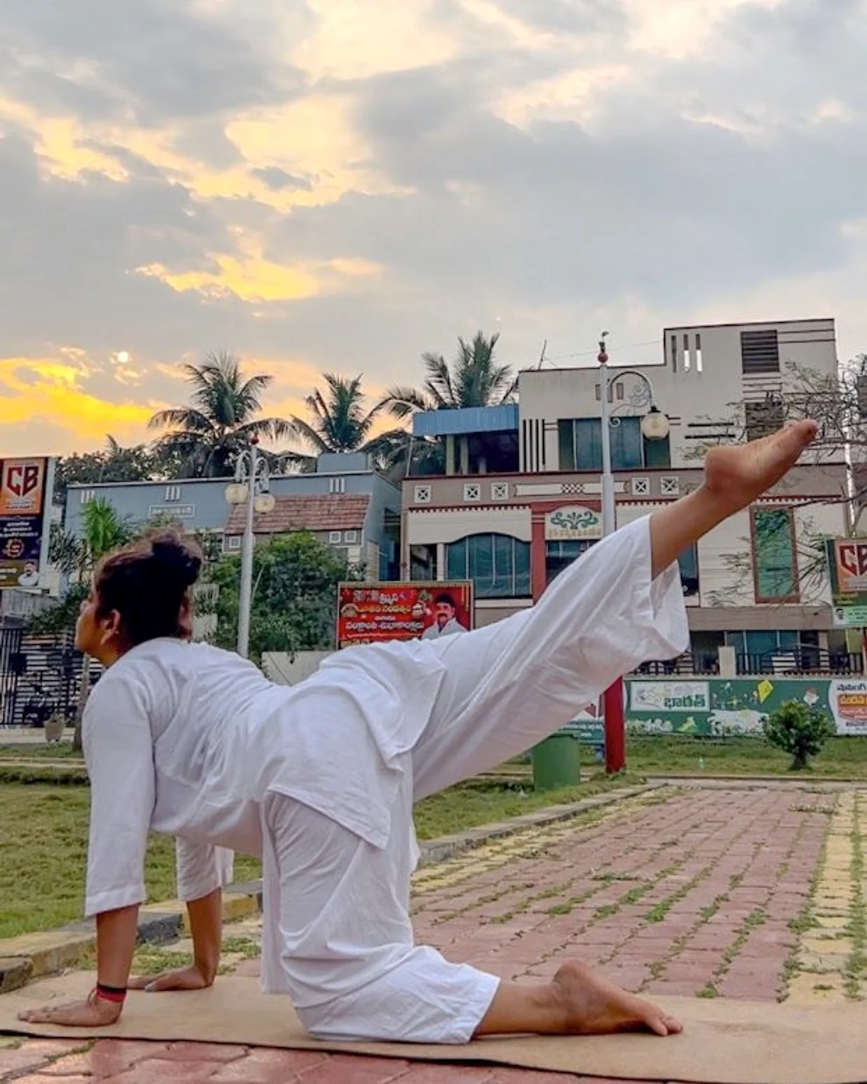A practitioner extends one leg in a balancing posture on a mat outdoors against a wide evening sky.