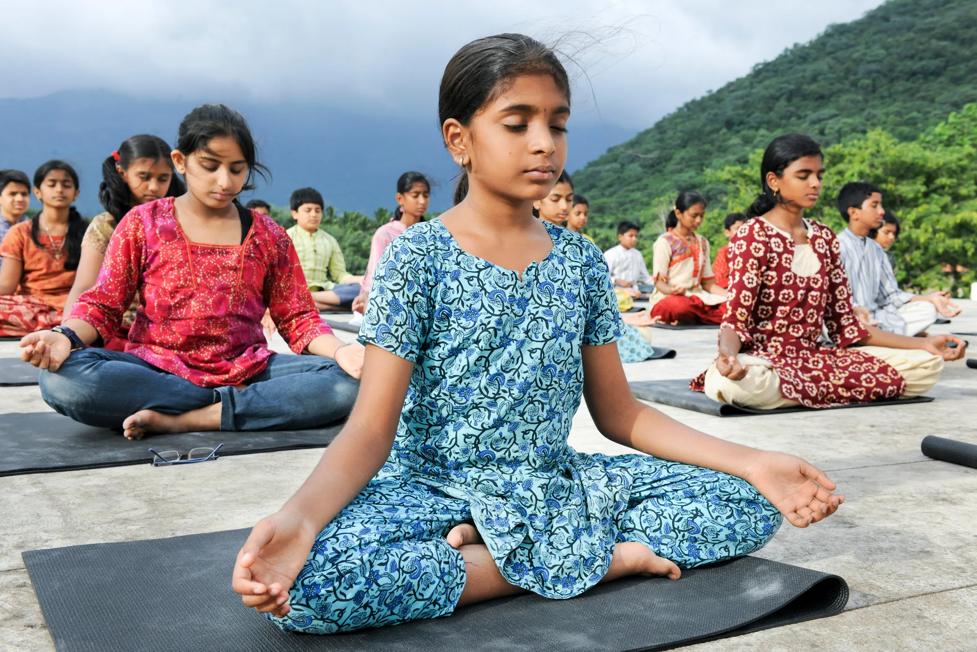 Children seated in quiet meditation on mats, mountains in the distance.