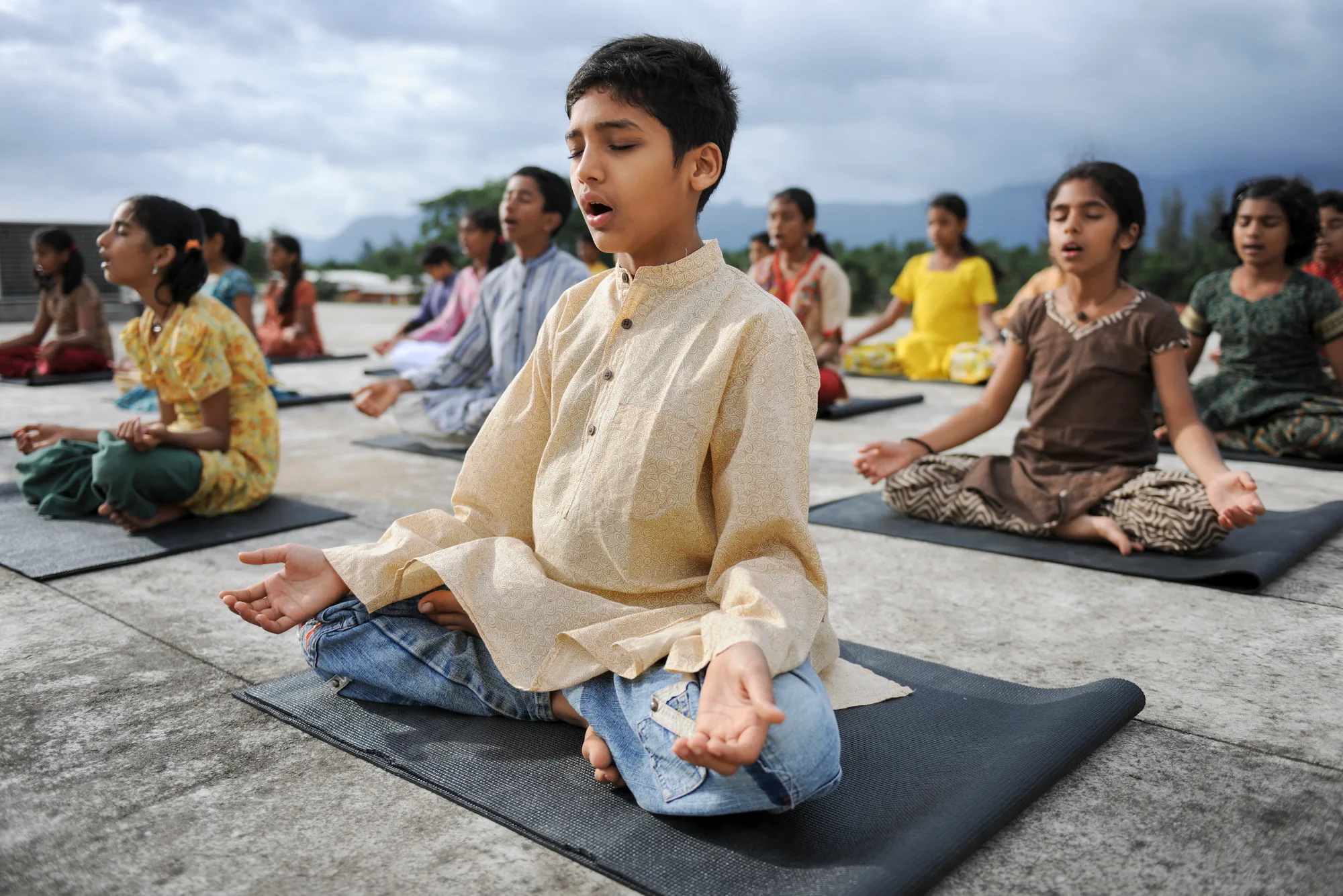 A group of young students sit in quiet meditation on mats, mountains softening behind them.